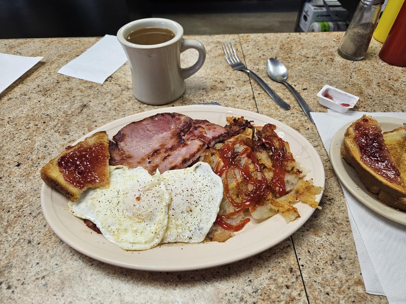 Ham steak, two fried eggs, hash browns and jam on toast with a mug of coffee on the counter