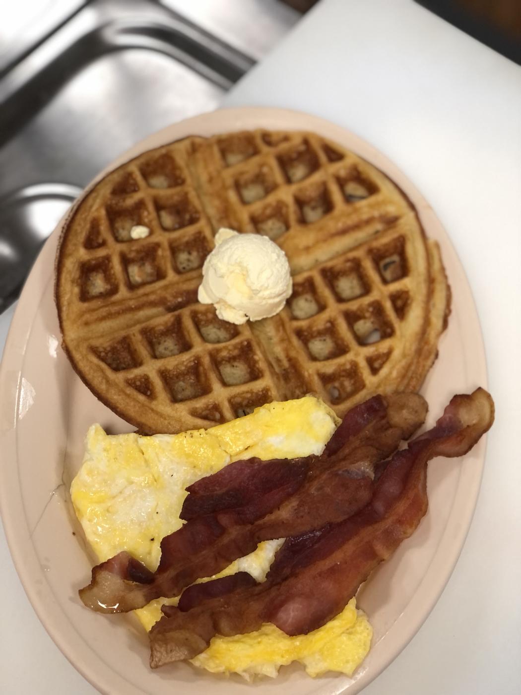 A waffle with a scoop of butter, scrambled eggs and bacon on a diner platter
